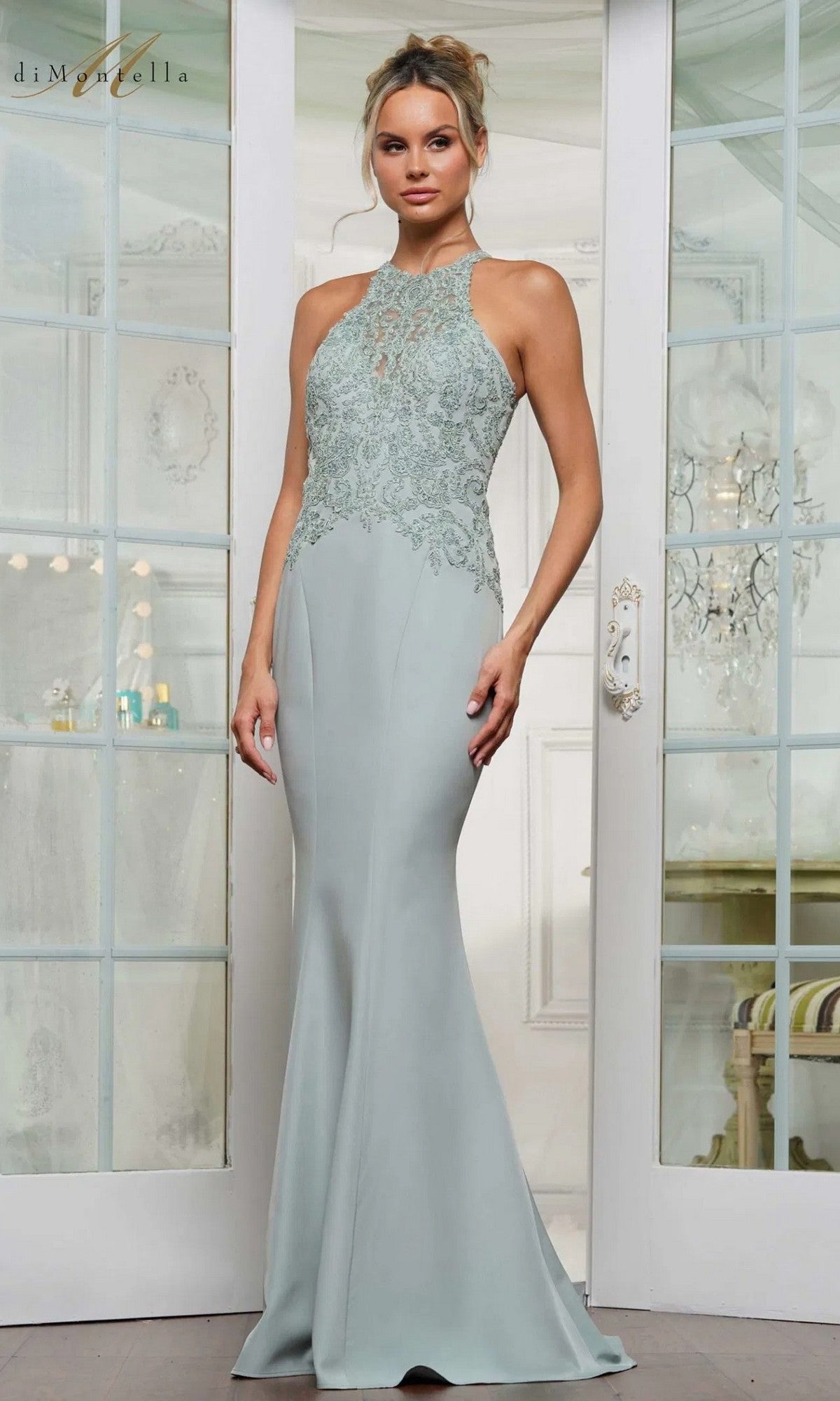 Wearing the Rina Di Montella RD3243 long formal dress in sage, featuring a lace-embroidered bodice and floor-length silhouette, a woman stands before glass-paneled doors, radiating timeless elegance.